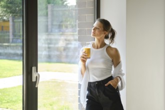 Cute woman stands thoughtfully by a large window, sipping a smoothies in a light-filled space. She