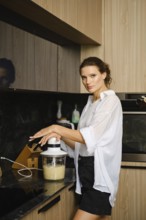 A woman stands at a kitchen counter, blending banana and sea buckthorn ingredients. Natural light