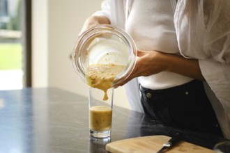 Pouring a creamy banana and sea buckthorn smoothie from a glass bowl into a tall glass. The setting