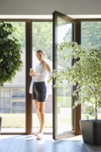 A woman stands gracefully at an open doorway, holding a smoothies. Sunlight floods the modern