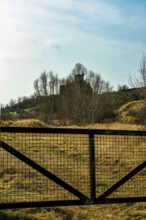 A picturesque view captures the ruins of a castle atop a hill, framed by a metal gate. The