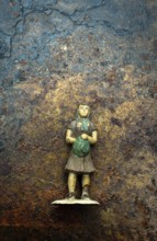 A small, weathered figurine depicting a girl in a dress, clutching a green bundle, stands on a