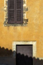 A textured yellow wall features a closed wooden window with shutters above a small door. Shadows
