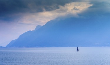 A lone sailboat navigates the tranquil waters of Lake Geneva surrounded by shadowy mountains. The