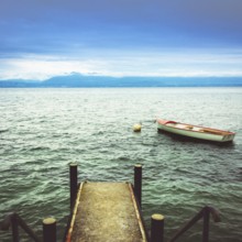 A wooden jetty reaches into the calm waters of Lake Geneva, where a small boat is moored. Mountains