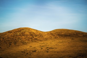 Two gentle hills rise from a golden landscape under a bright blue sky. Wispy clouds streak across