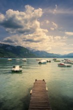 A wooden pontoon extends over the tranquil waters of Annecy Lake, surrounded by mountains and