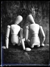 Two wooden figures are positioned closely together, facing away from the viewer. They sit on a
