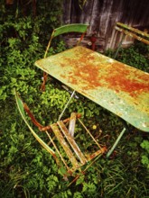 A rusty green table and a broken chair lay forgotten in thick, overgrown grass, evoking a sense of