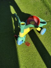 A vibrant, multi-colored tricycle sits on lush, green artificial grass, casting a shadow in bright