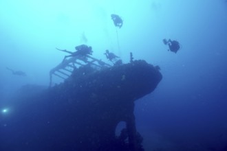Divers circle the silhouette of an overgrown shipwreck in the blue water. Dive site wreck Le Grecq,