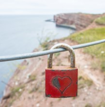 Red padlock with engraved heart symbol on a wire rope in front of the steep coast, red cliff of