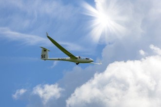 A glider flies through a blue sky with white clouds and bright sun, Baden-Württemberg, Germany