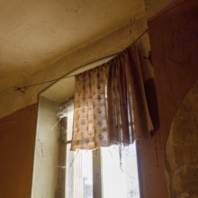 Sunlight streams through a dusty window adorned with worn curtains, highlighting the cobwebs in a