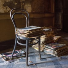 An old chair is perched beside a towering stack of newspapers, both covered in dust, in a dimly lit