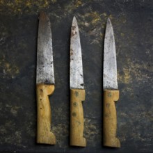 Three vintage kitchen knives lie on a dark, textured surface, their worn blades reflecting use,
