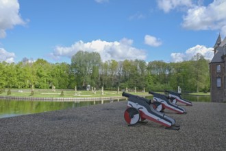 Small cannons at the edge of a park with a pond and sculptures, under a blue sky with clouds,