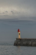 A lighthouse stands on a pier and casts its light over calm waters at dusk. The sky is filled with