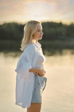 A young woman stands by a calm lake during sunset, feeling the gentle breeze. Her casual outfit