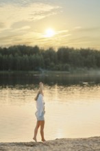 A woman stands on the sandy shore of a tranquil lake, raising her hand joyfully towards the setting