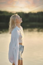 A woman stands by the calm lake, feeling the gentle breeze as she admires the colorful sunset sky.