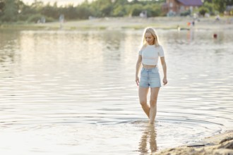 A young woman in a white crop top and denim shorts steps delicately into the clear water of a beach