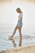 A woman stands in shallow water at a beach during golden hour, playfully splashing around with her