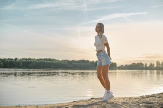 A young woman walks leisurely along the sandy lakeshore as the sun sets behind the trees. She wears