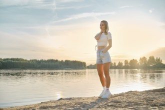 A young woman relaxing on the sandy beach near a calm river at sunset. She wears a casual outfit