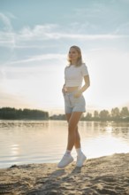 A young woman walks on tiptoes on a sandy shore as the sun sets over the lake. Her casual outfit