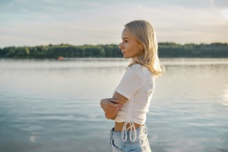 A blonde woman stands in profile by a calm lake, gazing thoughtfully into the distance. The sun