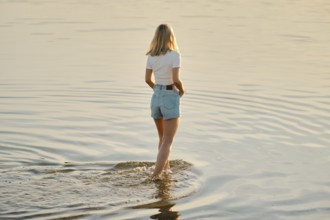 A woman with blonde hair wades through calm, shallow water as the sun sets behind her, creating a