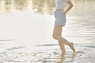 Unrecognizable woman stands in shallow water, playfully splashing while enjoying a serene sunset at