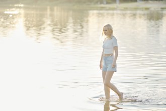 A young woman with light blonde hair walks barefoot in shallow water at sunset. The serene lake