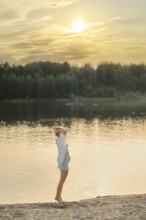 A woman stands at the edge of a tranquil lake during sunset, gazing across the water. Warm golden