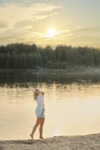 A barefoot woman walks along the sandy shore of the forest lake. She set her face to the warm rays