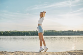 A young woman walking along a tranquil lake at sunset, dressed in casual summer attire. She