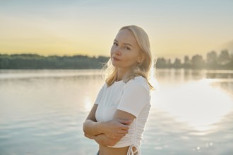 Cute woman stands thoughtfully by the lake, gazing into the distance as the sun sets behind her.