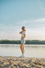 A young woman stands on the sandy shore of a tranquil lake, gazing at the water while wearing