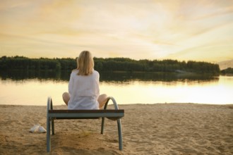 A woman with blond hair sits on a beach bench, gazing at the serene lake at sunset, surrounded by