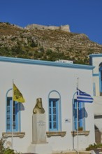 Bust in front of a white building with blue windows, Greek flag and yellow flag in the foreground,