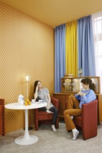 Two friends are sitting comfortably in a living room with bold wallpaper and bright curtains. They