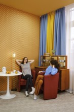 Two friends sit in elegant chairs within a warmly decorated room, engaged in a casual conversation