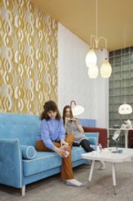 Two friends enjoy a cozy moment in a modern lounge, sitting on a colorful sofa. One woman removes