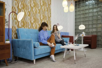 Two women are seated on a blue sofa in a living room, enjoying each other's company while engaging