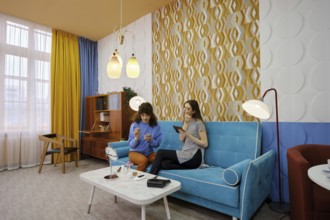 Two friends sit on a blue couch, engaged in conversation, while doing make-up and enjoying each