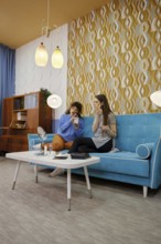 Two women sit on a bright blue couch in a stylish living room, laughing and applying beauty