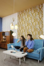 Two women remove makeup from their faces sitting at home on a blue couch in a cozy living room,