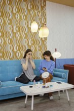 Two friends are sitting comfortably on a blue couch in a modern salon, engaging in a makeup session