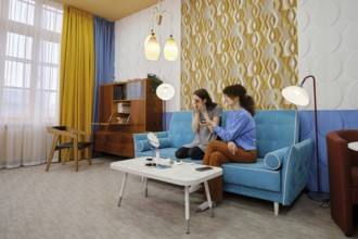 In a bright living room adorned with curtains, two friends sit on a blue sofa, chatting and